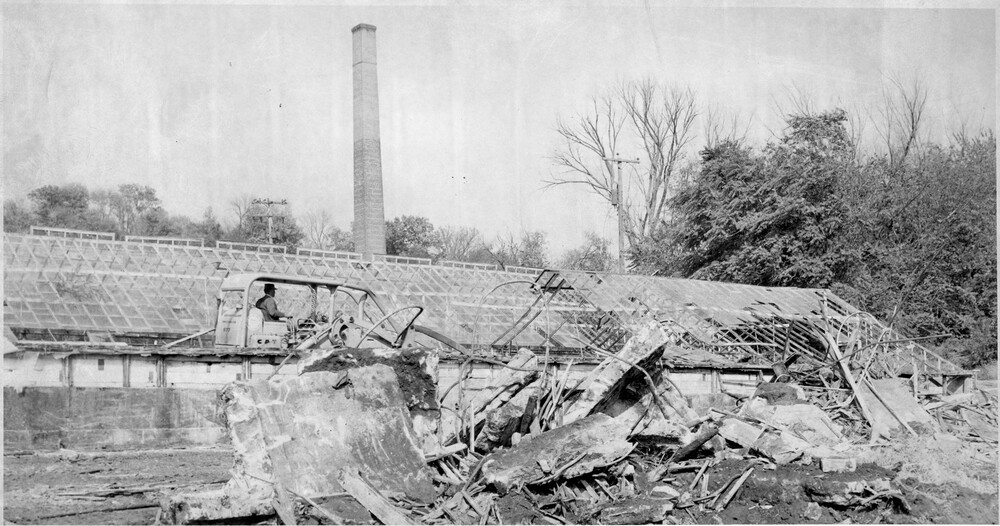 Bulldozer and wreckage from demolition in front of a greenhouse and brick chimney tower