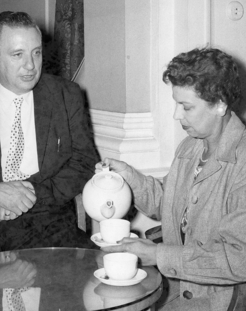 A woman sitting at a table pouring tea into a cup as a man watches. 