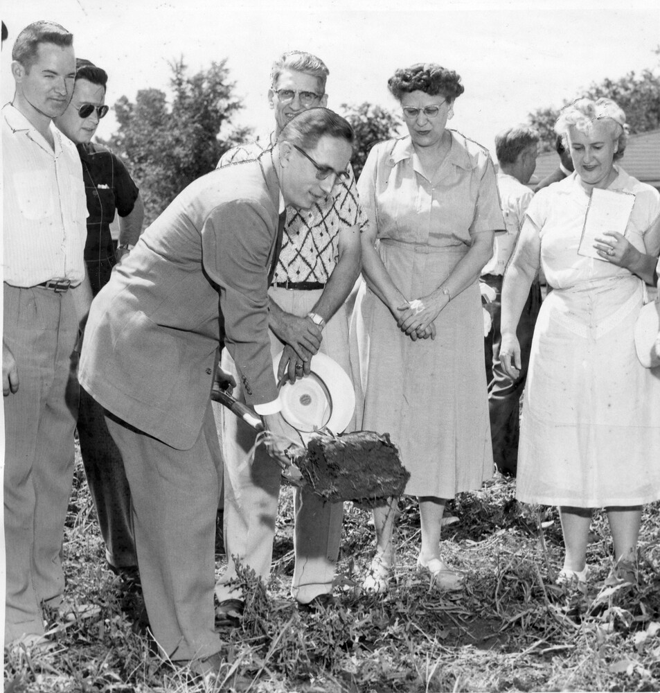 Man in suit lifts shovel of dirt, with five other people watching.
