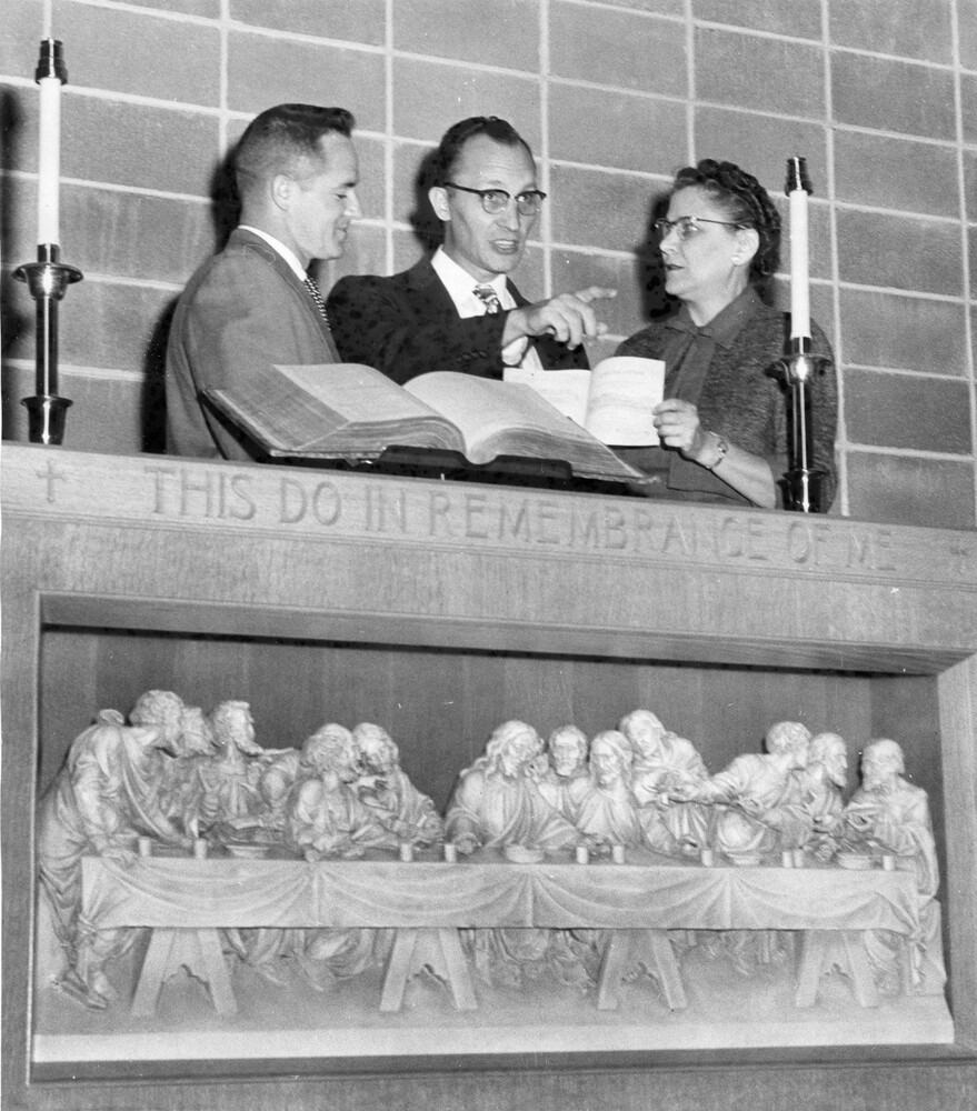 A woman and two men stand behind an altar faced with carving of the Last Supper; on altar are open book and two candlesticks.