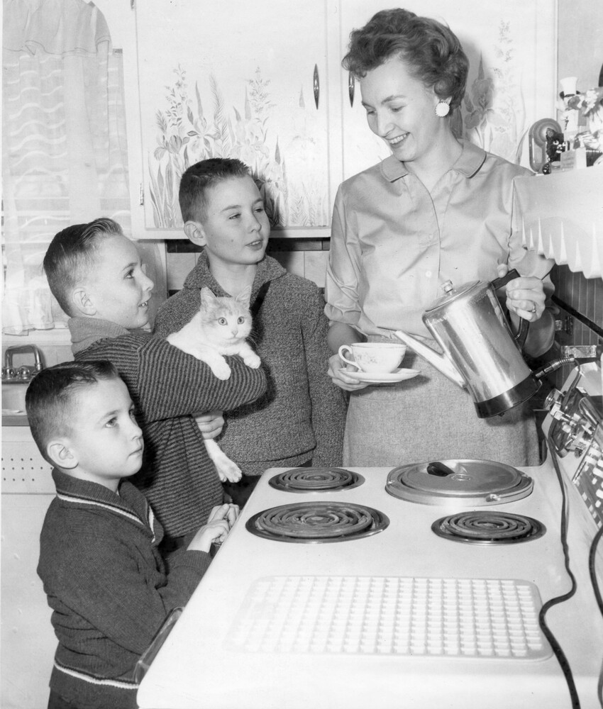 A woman holding a cup and saucer and an electric coffee pot stands at a stove with three young boys and a cat