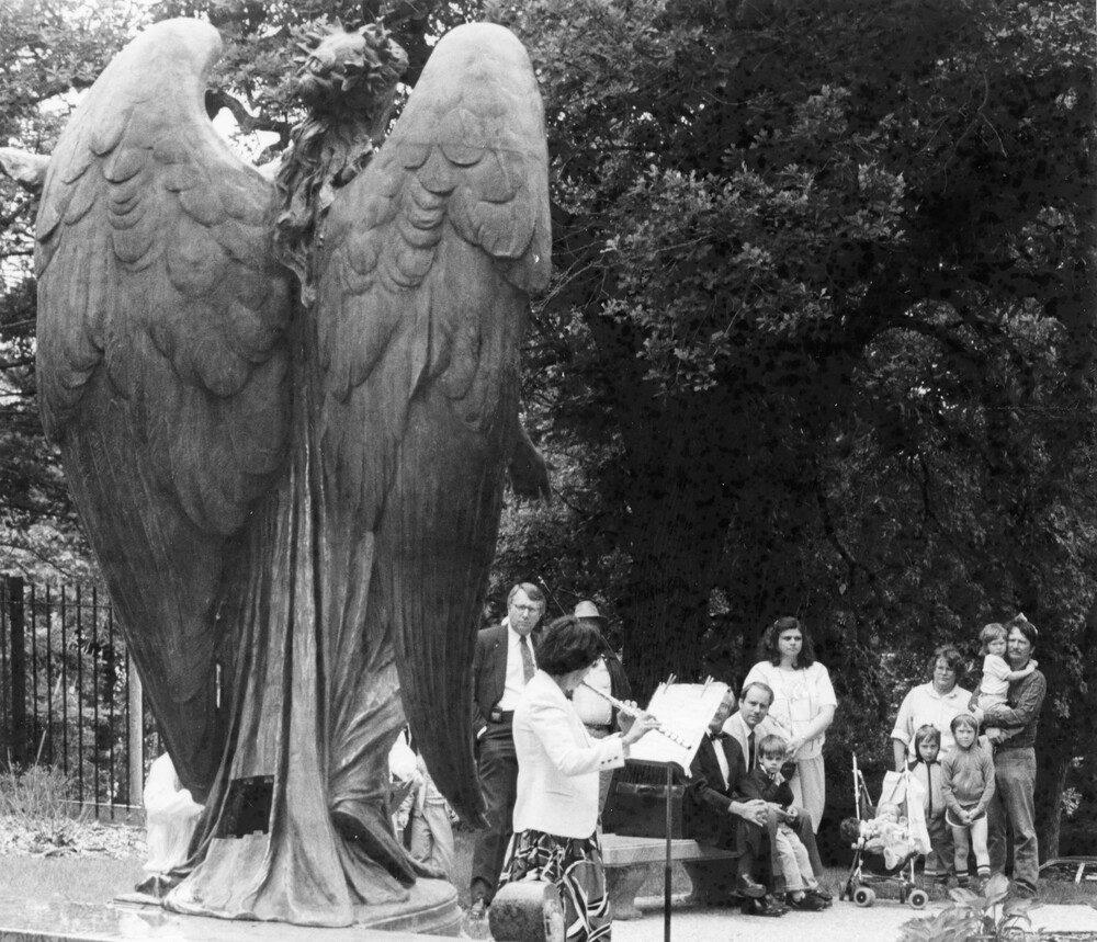 back of a large outdoor statue of an angel, facing flautist playing before audience