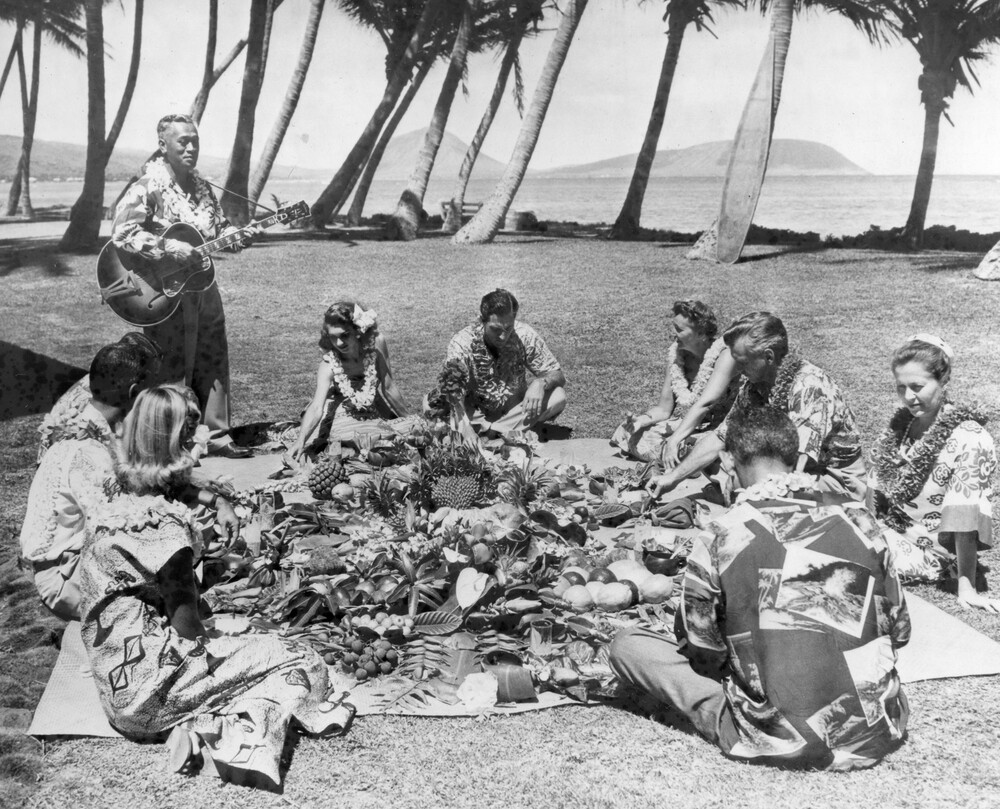 Eight people seated on the ground at a luau and a man playing a guitar.