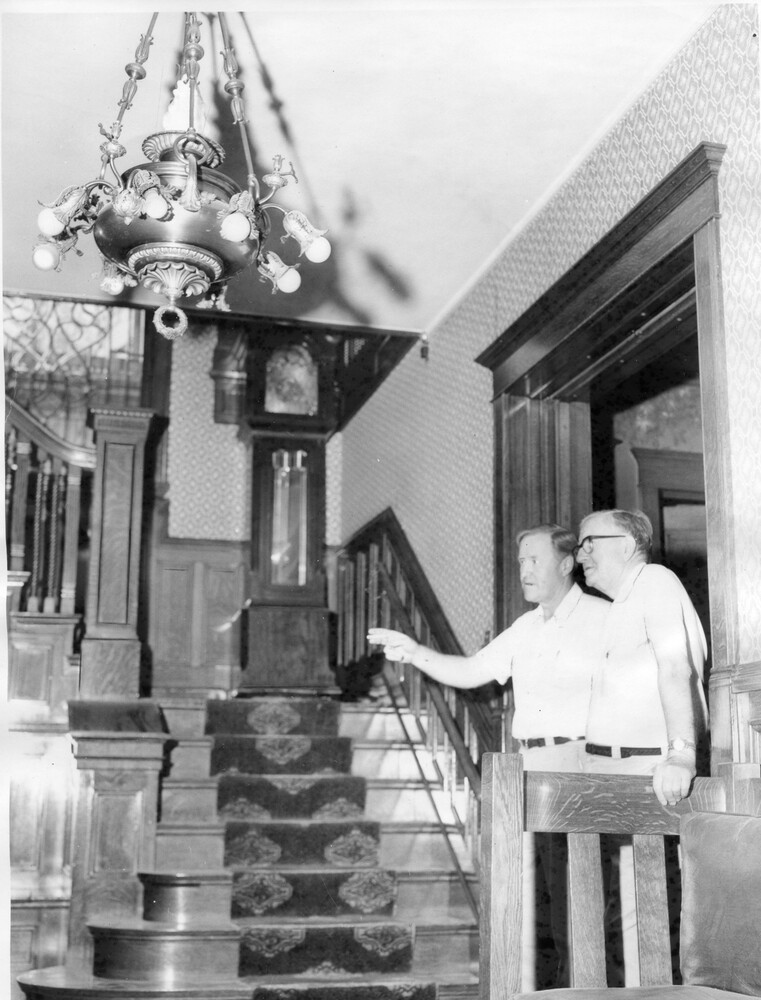 Photo of two men standing at the foot of large wooden staircase, with grandfather clock on the landing and chandelier overhead