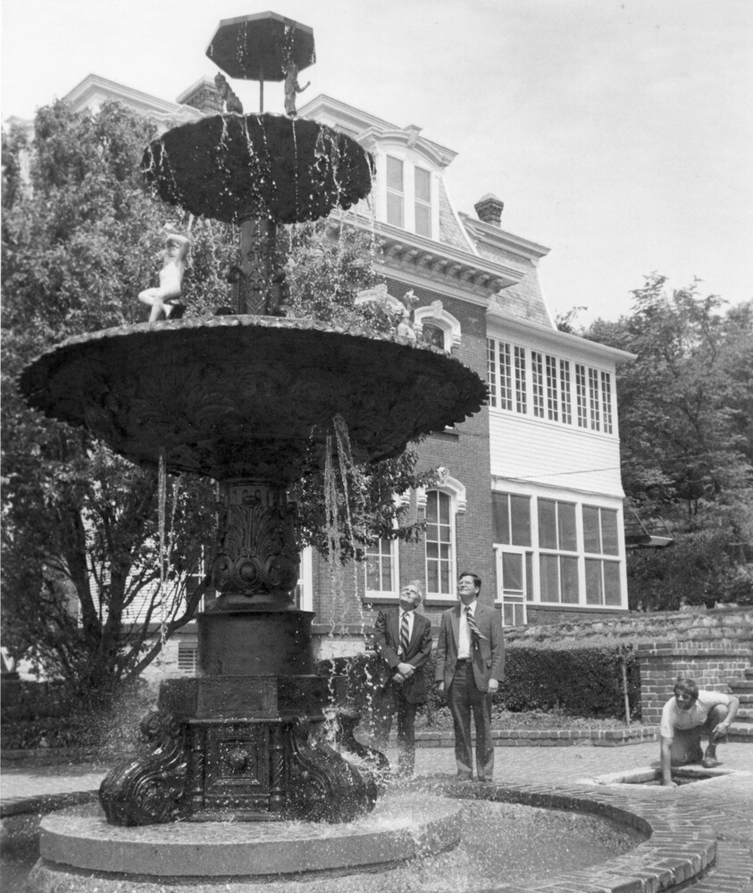 Two men standing by outdoor fountain, with another man turning on the water.