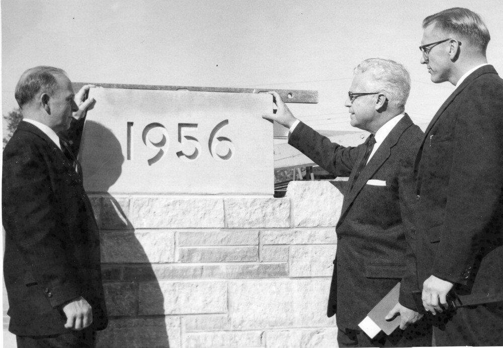 Three men in suits gather around a cornerstone reading "1956".