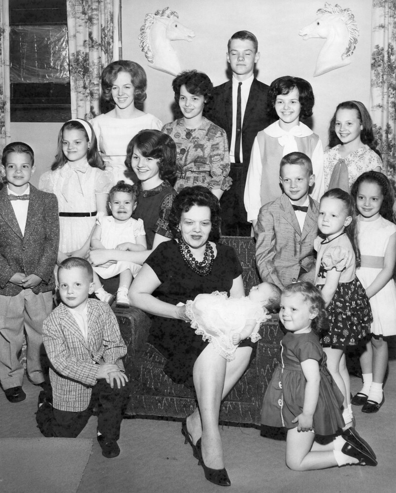Woman seated, holding a baby, and surround by 14 other children