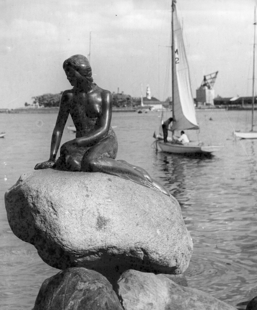 Mermaid statue on a rock in a harbor. Sailboat in background.