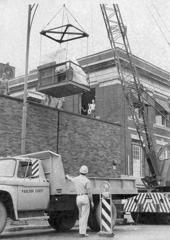 A large piece of machinery swings on a crane as a man standing next to a truck looks on. 