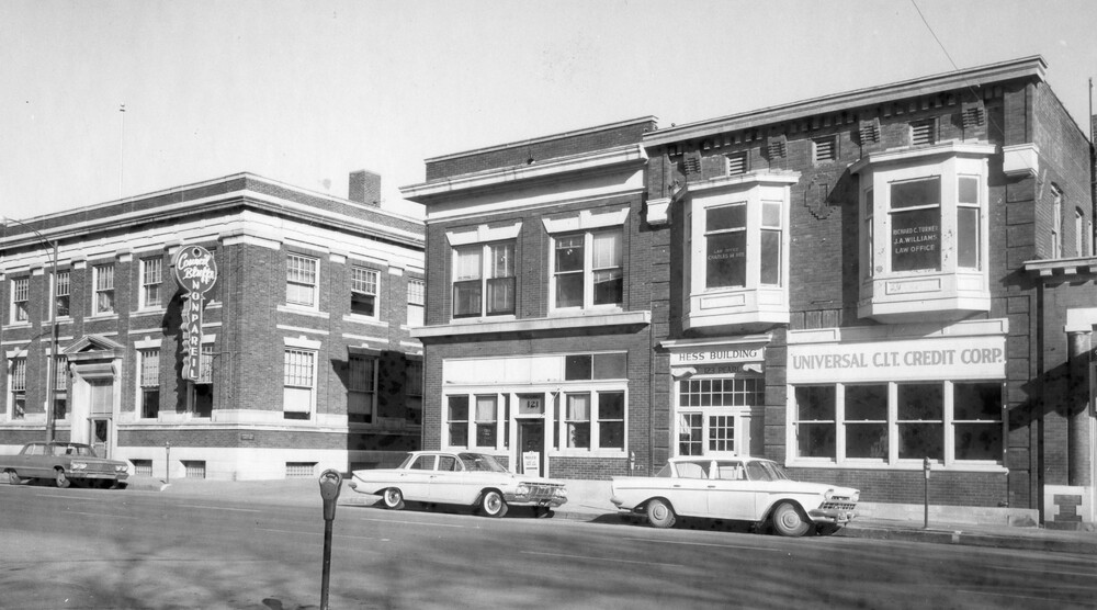 Cars parked on a street in front of three brick buildings.