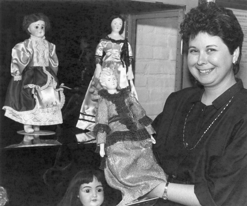 Woman holding antique doll, next to shelves with 3 more dolls.