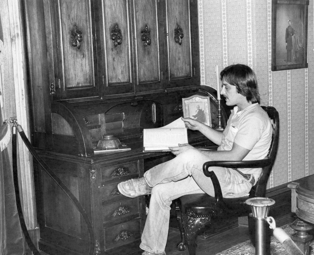 Man sits at rolltop desk, reading a book.