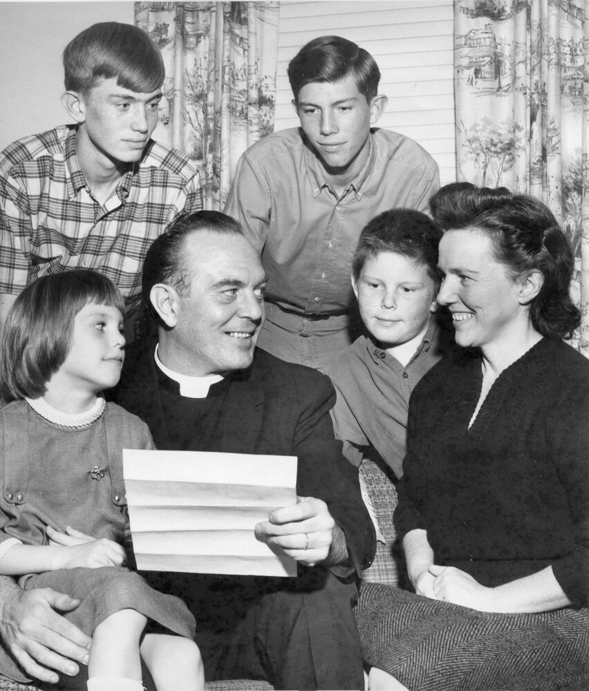 Family gathered around a sofa; father, wearing clerical collar, holds piece of paper that others are looking at.