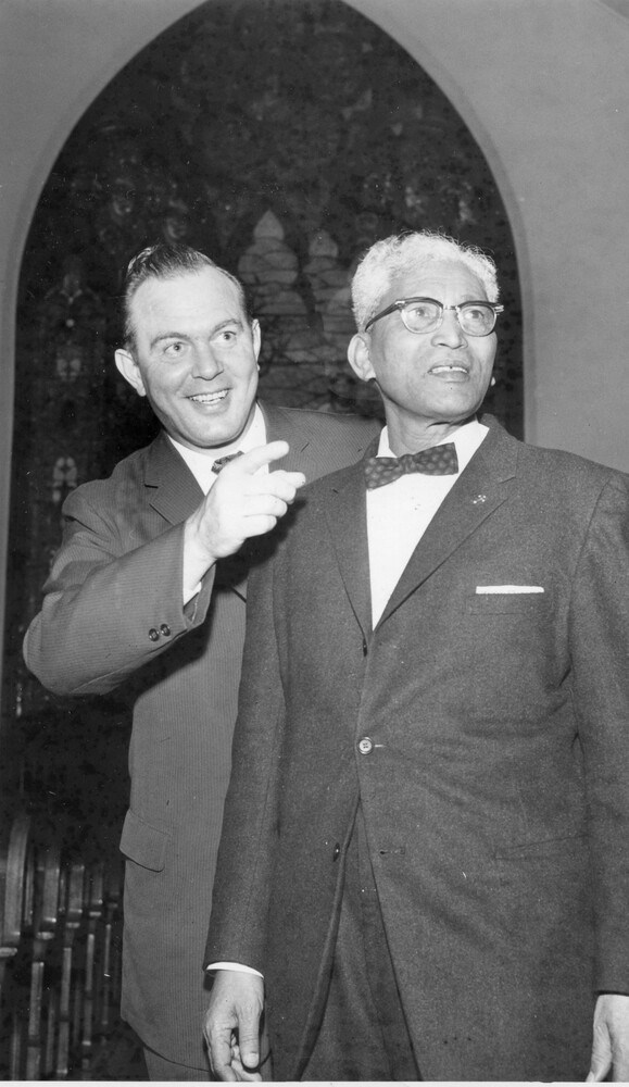 Two men in suits stand together, with one of the men pointing in front of them