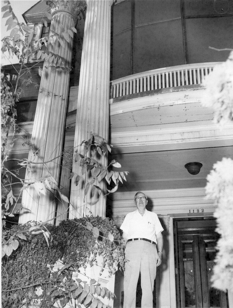 Photo of a man in shirtsleeves standing by columns of the mansion's veranda.