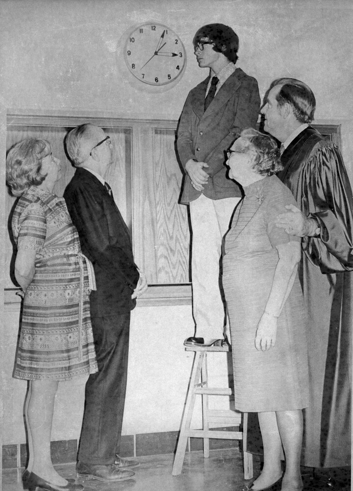 A young man stands on stepladder next to a clock, four people look on.
