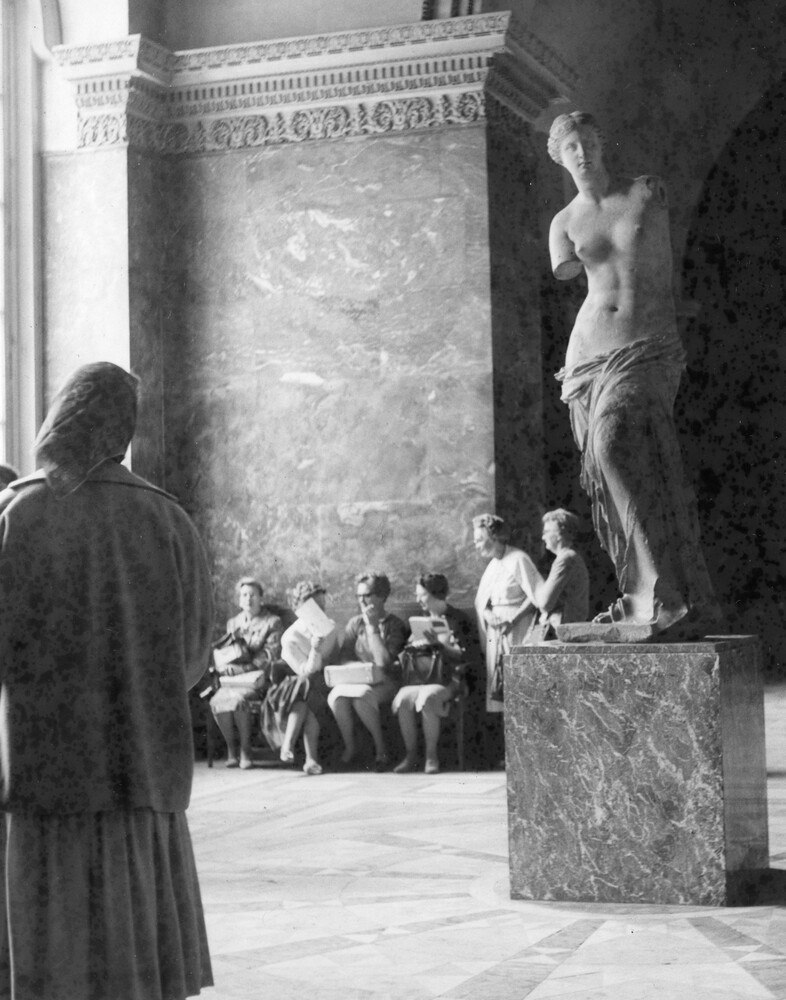 A group of women sitting on a bench in a sculpture gallery. 
