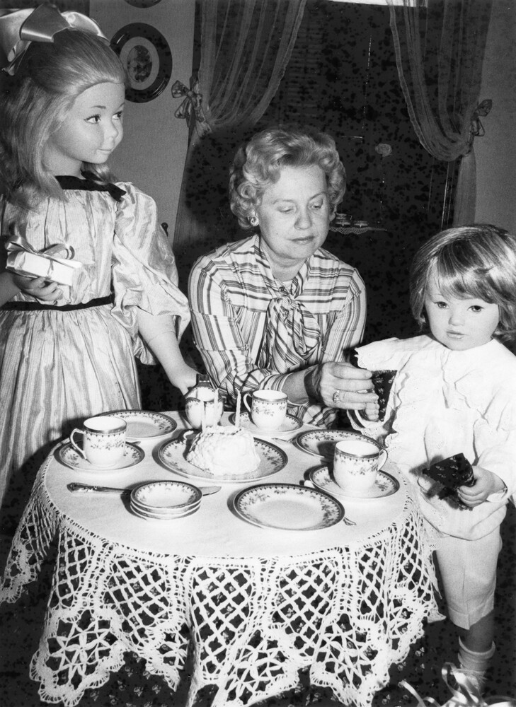 Woman sitting at small table set for tea, flanked by 2 standing mannequins of children.