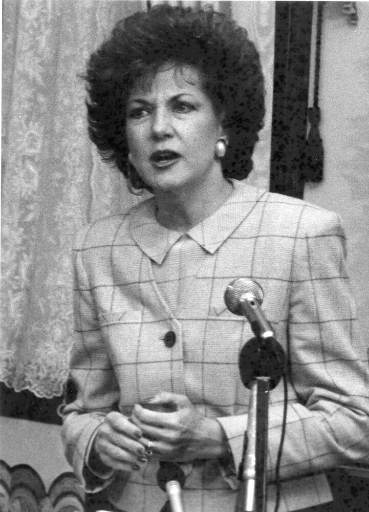 Woman in a suit stands behind microphones, speaking