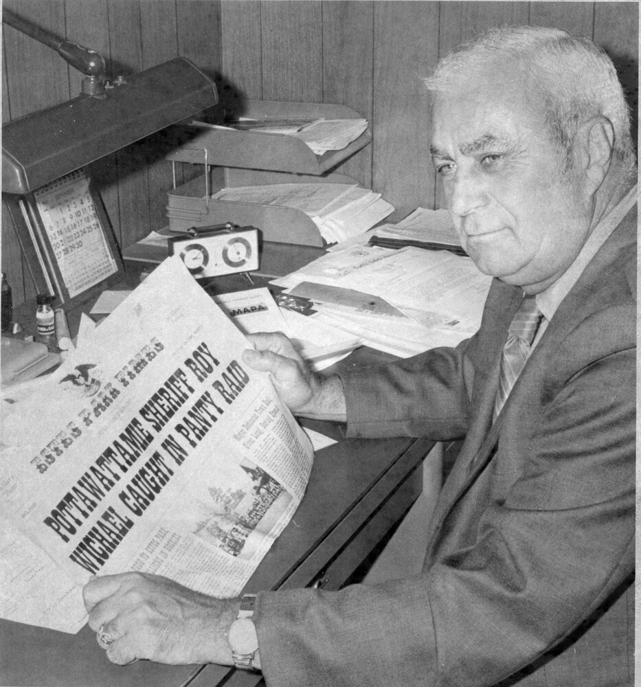Man in a suit sits at a desk, holding a newspaper showing a large headline