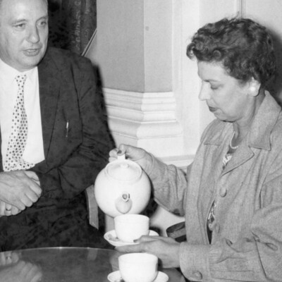 A woman sitting at a table pouring tea into a cup as a man watches. 