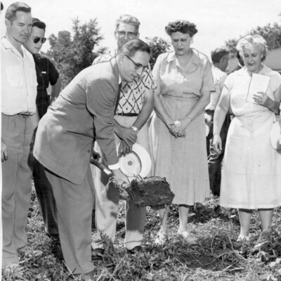 Man in suit lifts shovel of dirt, with five other people watching.