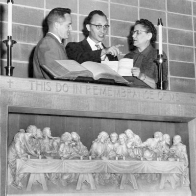 A woman and two men stand behind an altar faced with carving of the Last Supper; on altar are open book and two candlesticks.