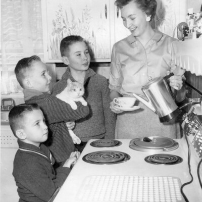 A woman holding a cup and saucer and an electric coffee pot stands at a stove with three young boys and a cat