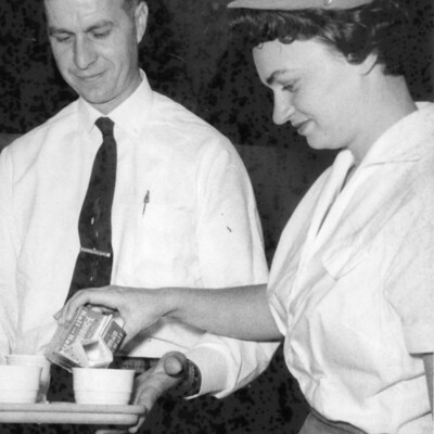Woman pouring cream into coffee cup held on a tray by a man.