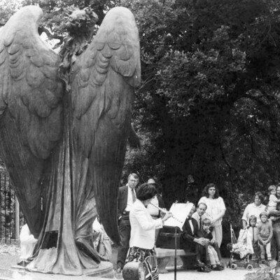 back of a large outdoor statue of an angel, facing flautist playing before audience