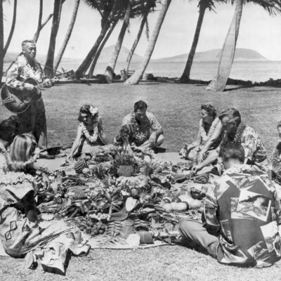 Eight people seated on the ground at a luau and a man playing a guitar.