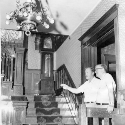Photo of two men standing at the foot of large wooden staircase, with grandfather clock on the landing and chandelier overhead