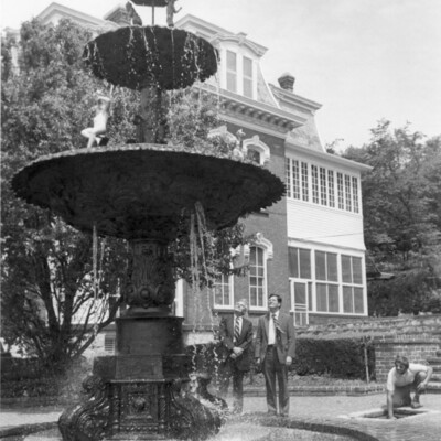 Two men standing by outdoor fountain, with another man turning on the water.