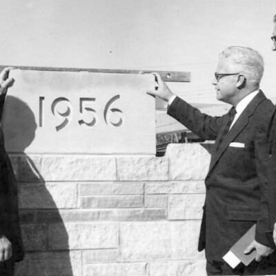 Three men in suits gather around a cornerstone reading "1956".