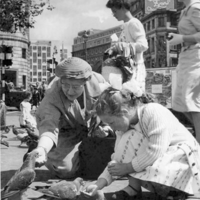 Two women crouching to feed pigeons with women in the background on a street.