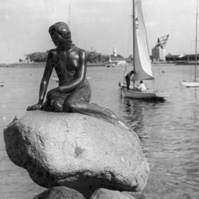 Mermaid statue on a rock in a harbor. Sailboat in background.