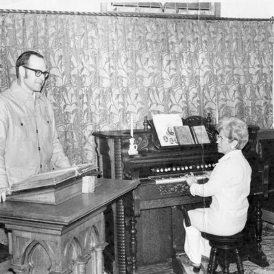 A man standing behind a pulpit and a woman playing an organ. 