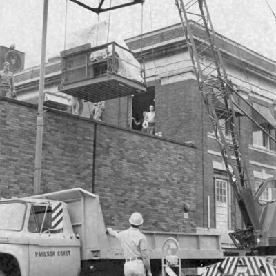 A large piece of machinery swings on a crane as a man standing next to a truck looks on. 