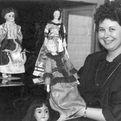 Woman holding antique doll, next to shelves with 3 more dolls.