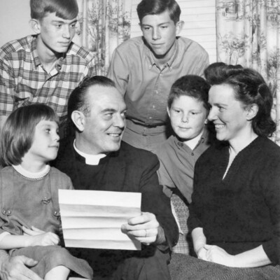 Family gathered around a sofa; father, wearing clerical collar, holds piece of paper that others are looking at.