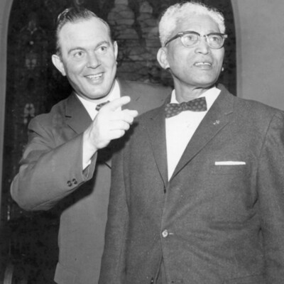 Two men in suits stand together, with one of the men pointing in front of them