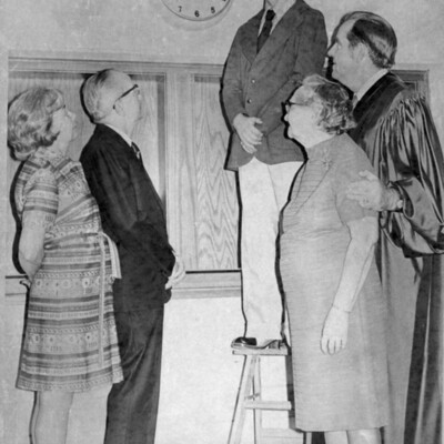 A young man stands on stepladder next to a clock, four people look on.