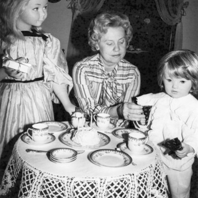 Woman sitting at small table set for tea, flanked by 2 standing mannequins of children.