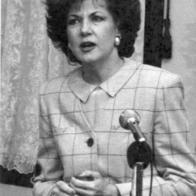 Woman in a suit stands behind microphones, speaking
