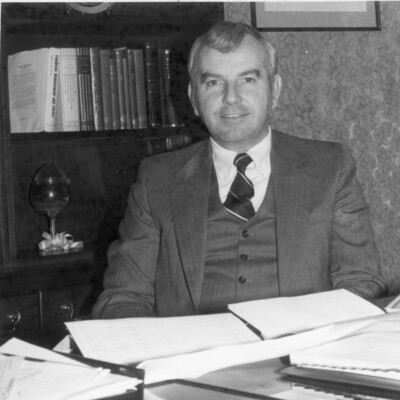 Man in suit in tie, seated behind desk with papers and open files