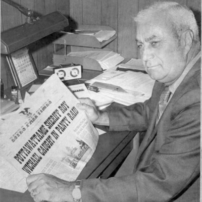 Man in a suit sits at a desk, holding a newspaper showing a large headline
